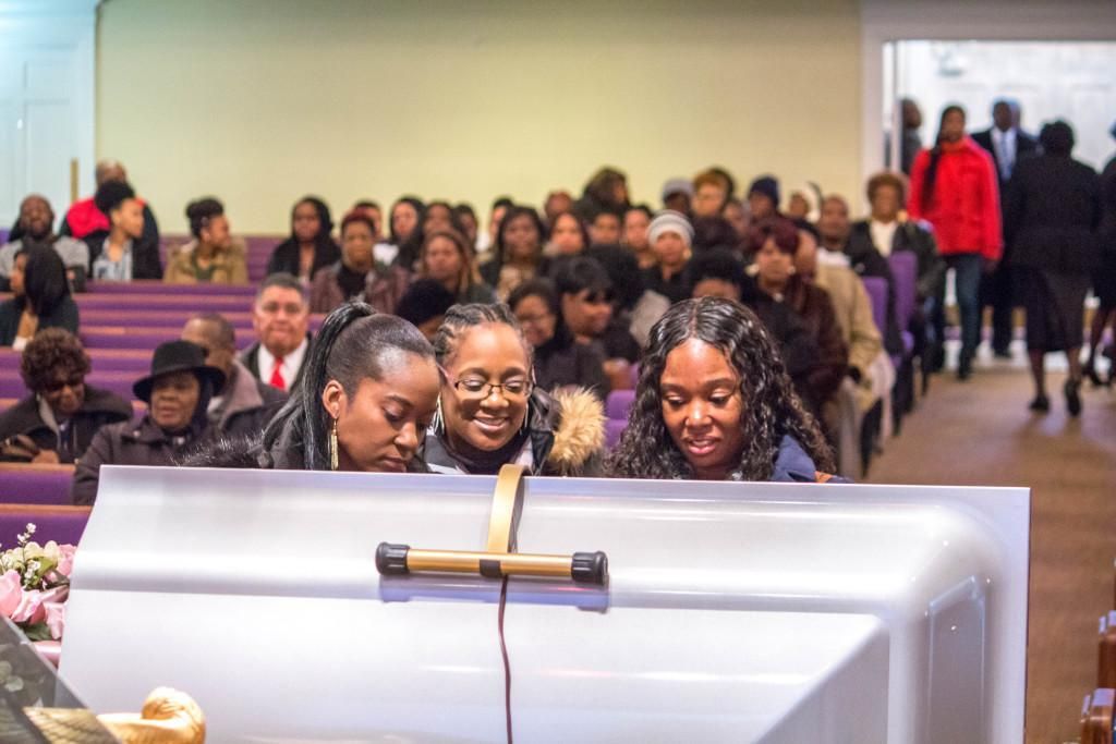 People gathered at a funeral service; three women look down at a closed silver casket.