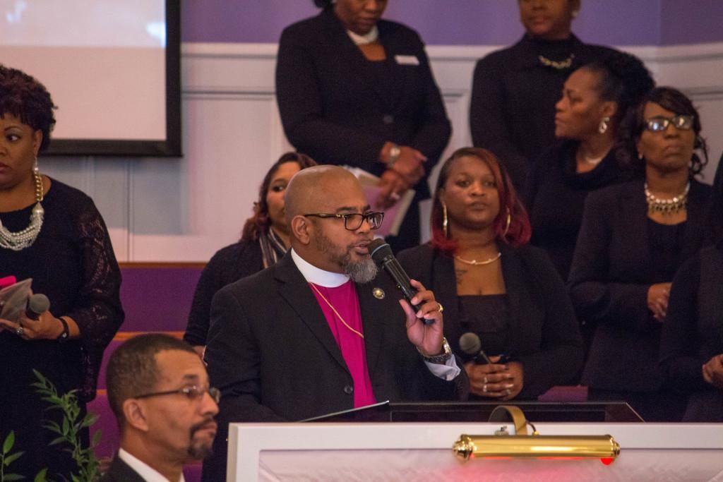 Man in black suit with mic speaks at a podium; people in formal attire behind him.