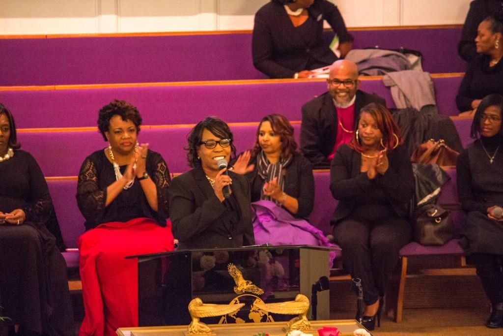 People clapping at a church service; one woman speaks into a microphone. Purple pews.