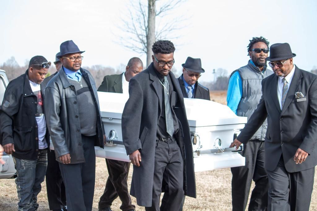 People carrying a white coffin at a funeral in an outdoor setting.