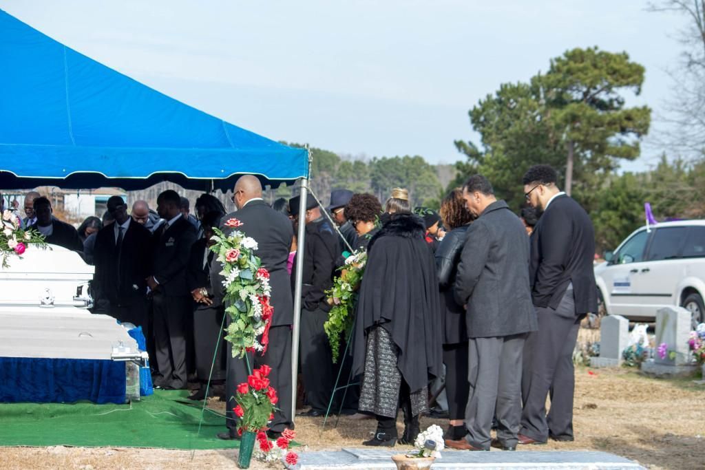 Mourners gather at a graveside funeral under a blue tent; casket and wreath visible.