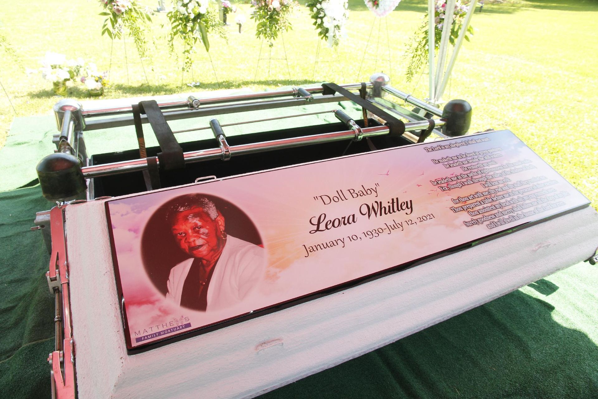 Casket with photo and text, set on green grass. Flowers above. A funeral service.