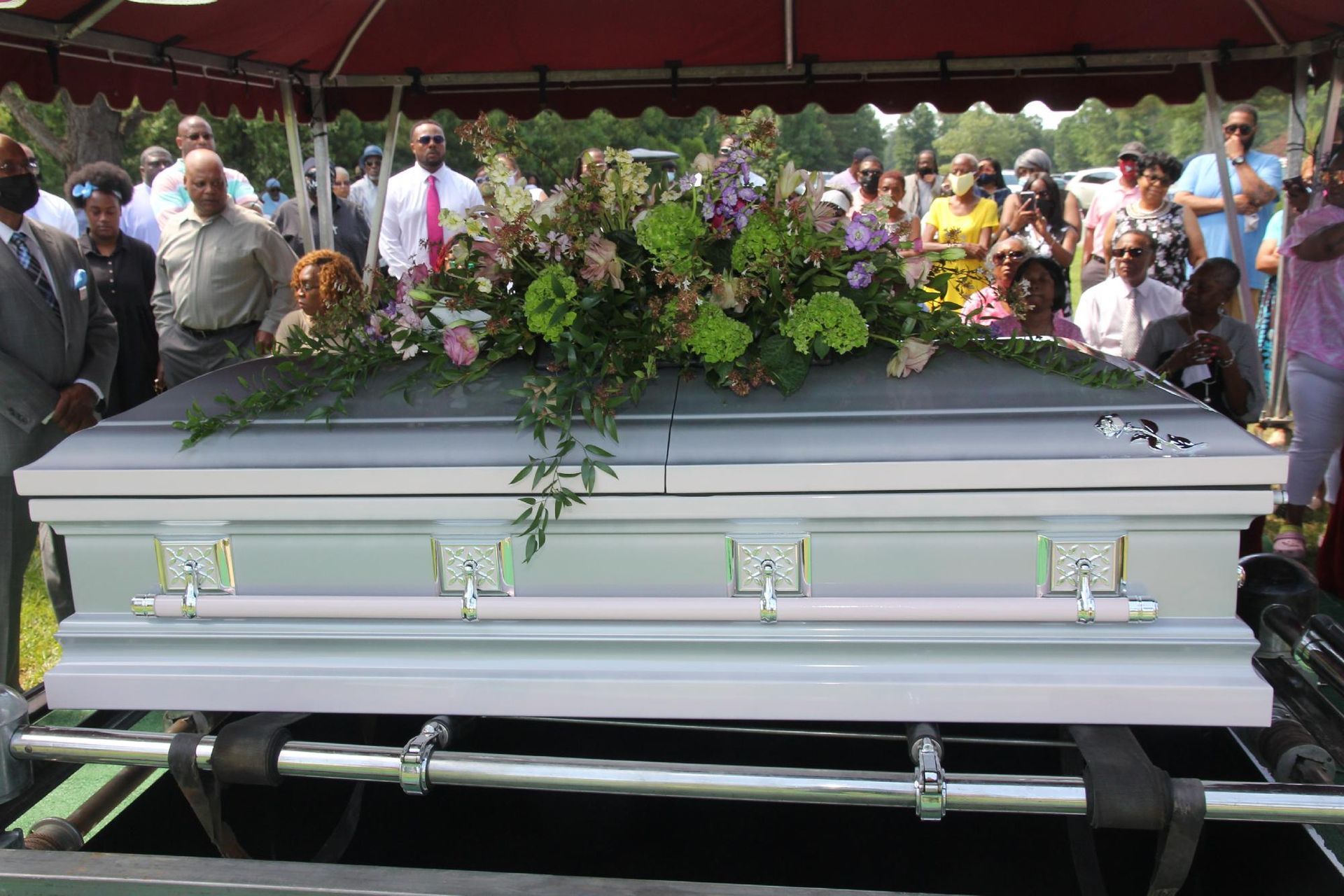 Light blue casket at a graveside service, adorned with floral arrangement, surrounded by mourners.