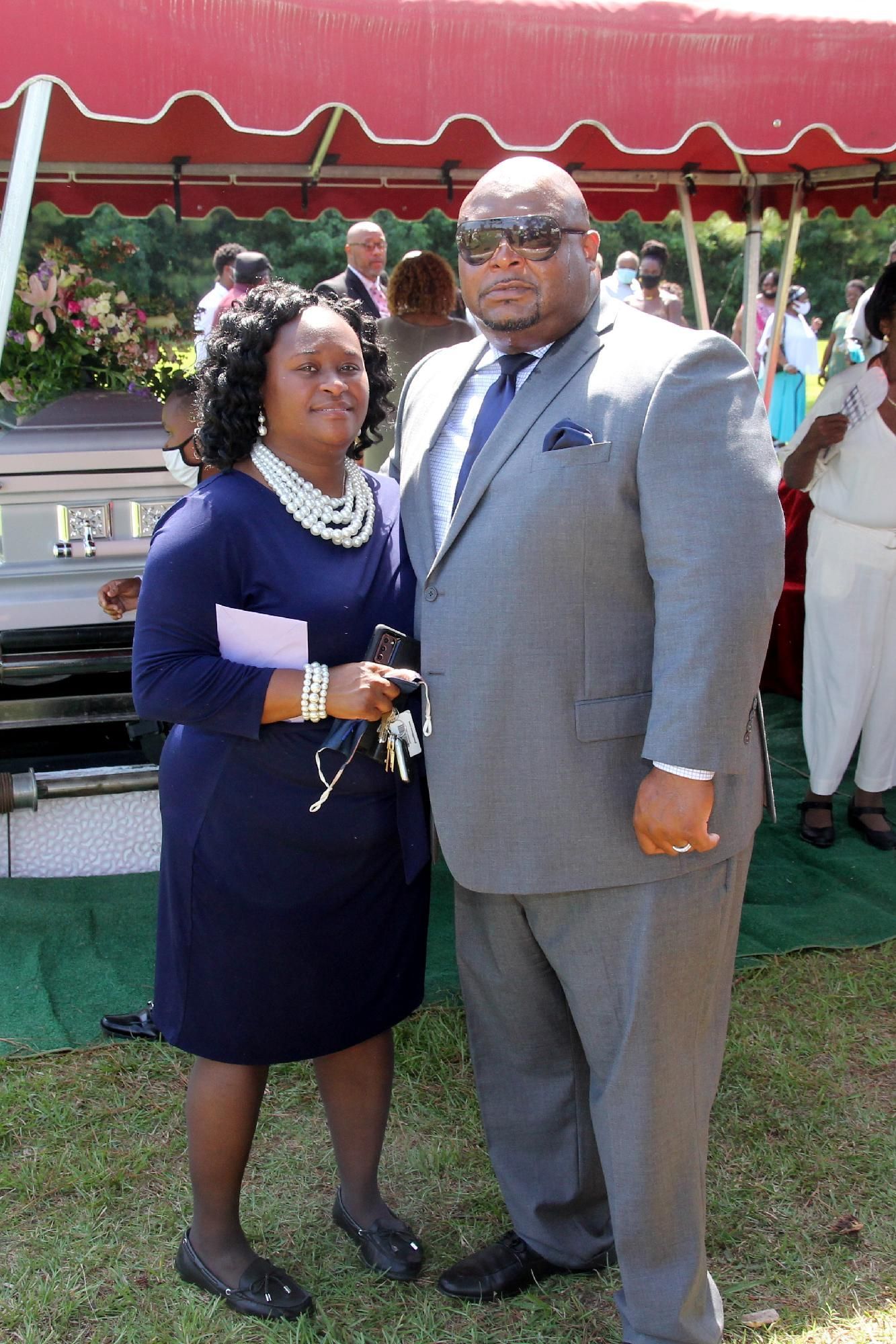 A couple poses near a casket at an outdoor funeral. Woman in blue dress, man in gray suit.