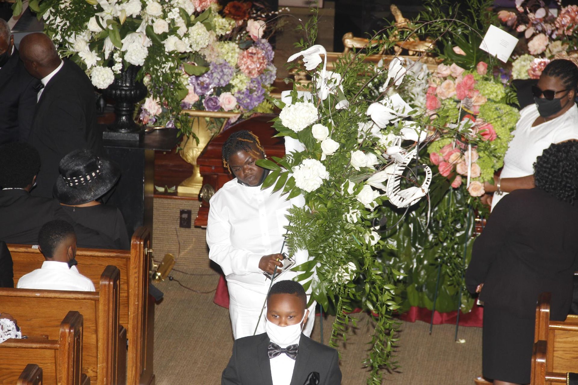 Funeral service with flowers and mourners, one person in white attire is praying.
