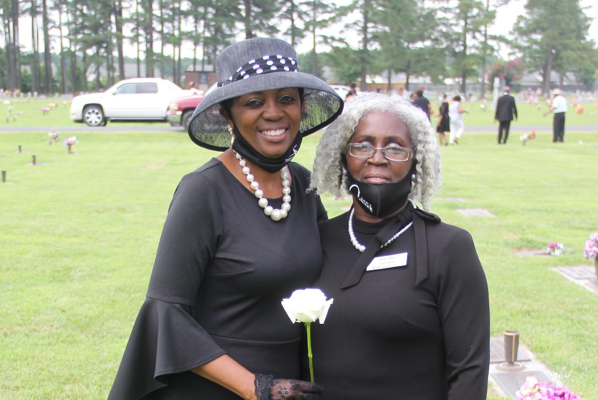 Two women in black at a cemetery, one holding a white rose, both wearing masks.