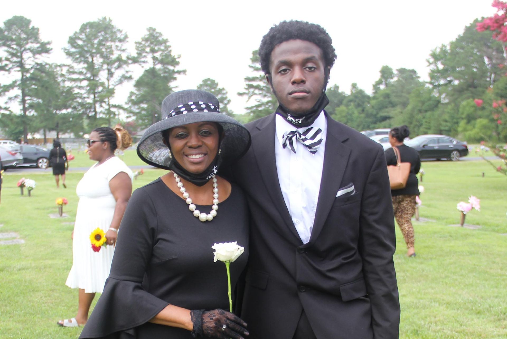 Woman and young person pose in cemetery. Woman wears black hat, dress, pearls, holds white rose.