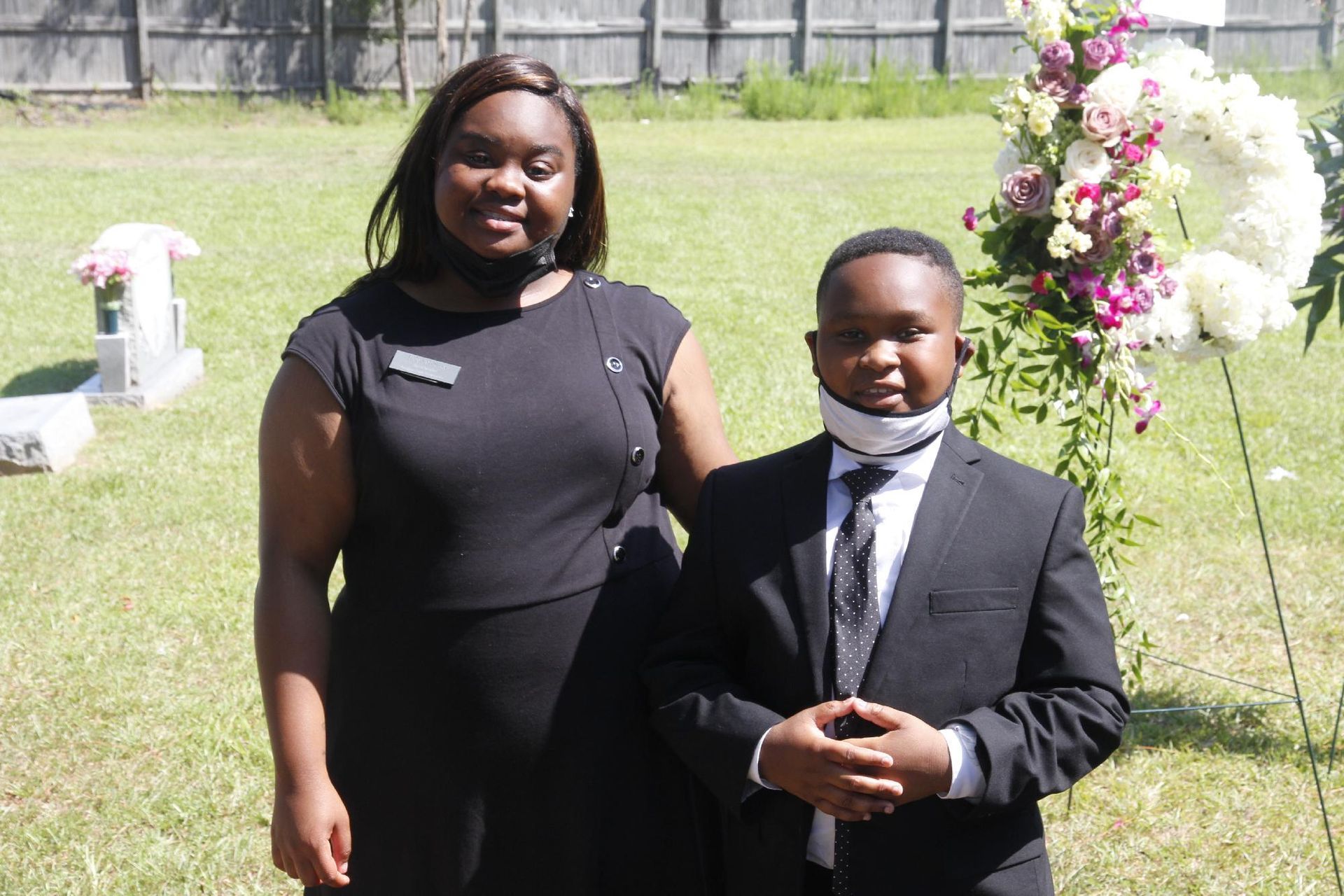 Woman and boy beside floral wreath at a gravesite; both wearing masks and dark attire.