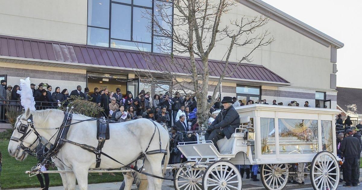 Horse-drawn carriage carrying a casket in front of a building; large crowd gathered outside.