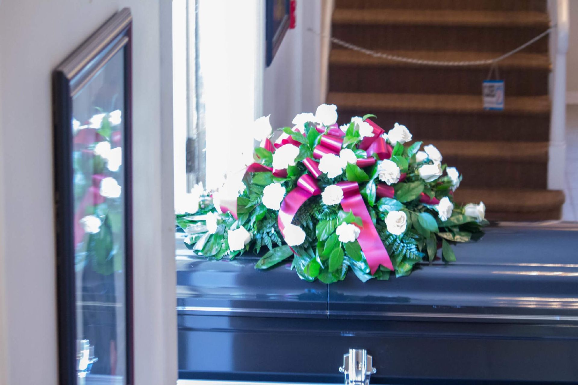 Floral arrangement of white roses and a burgundy ribbon on a dark coffin, indoors.