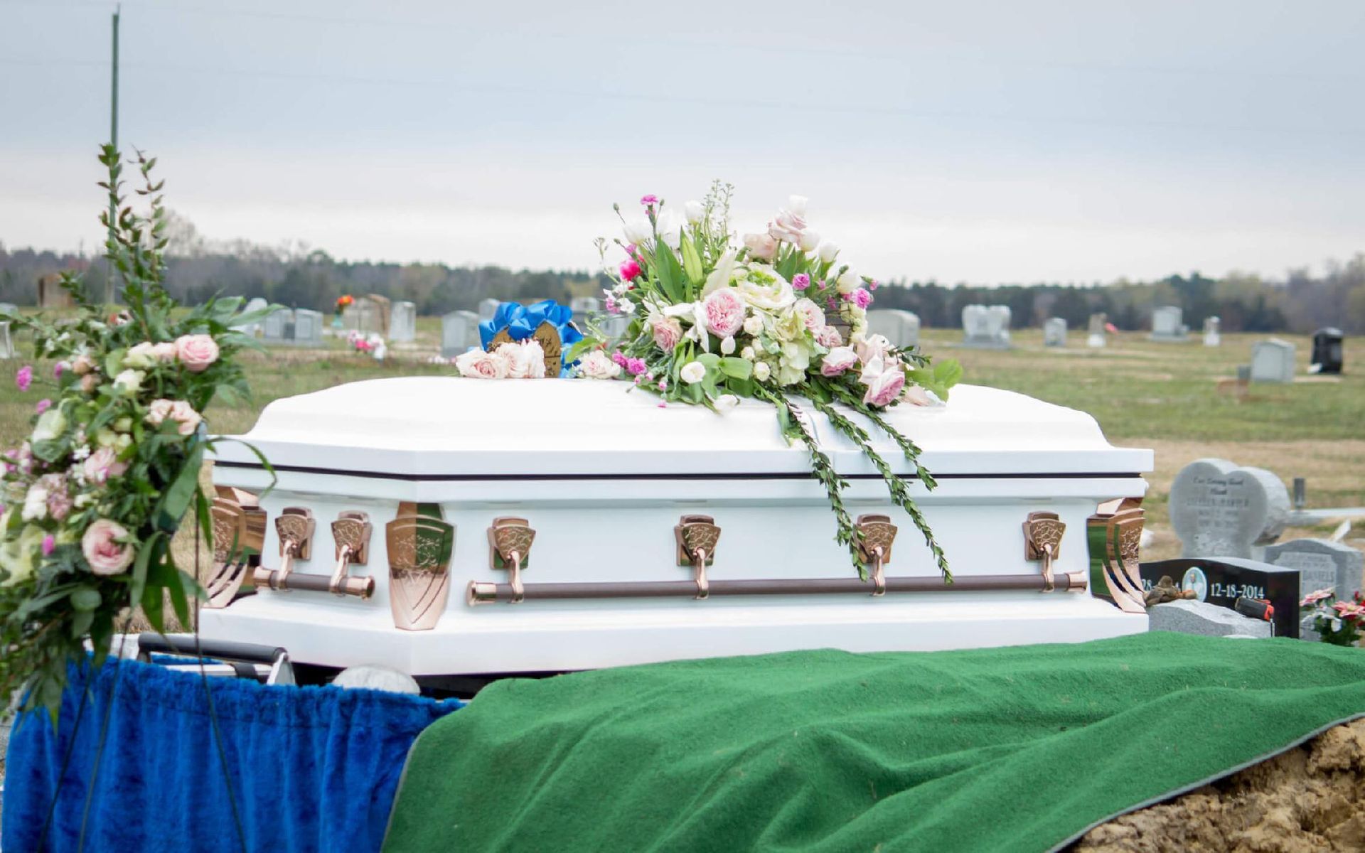 White casket with floral arrangement at a cemetery, blue and green cloth draping.