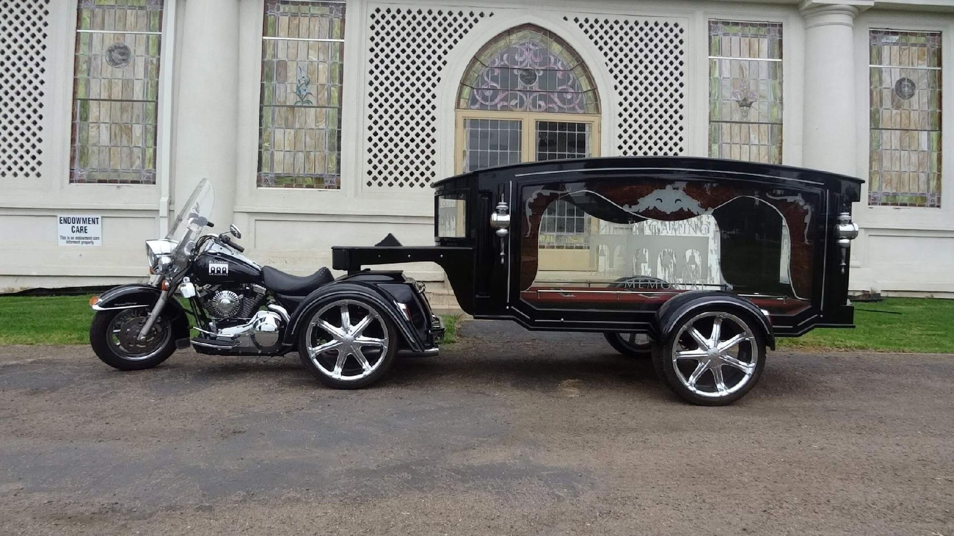 Black motorcycle towing a hearse trailer in front of a white building with arched windows.