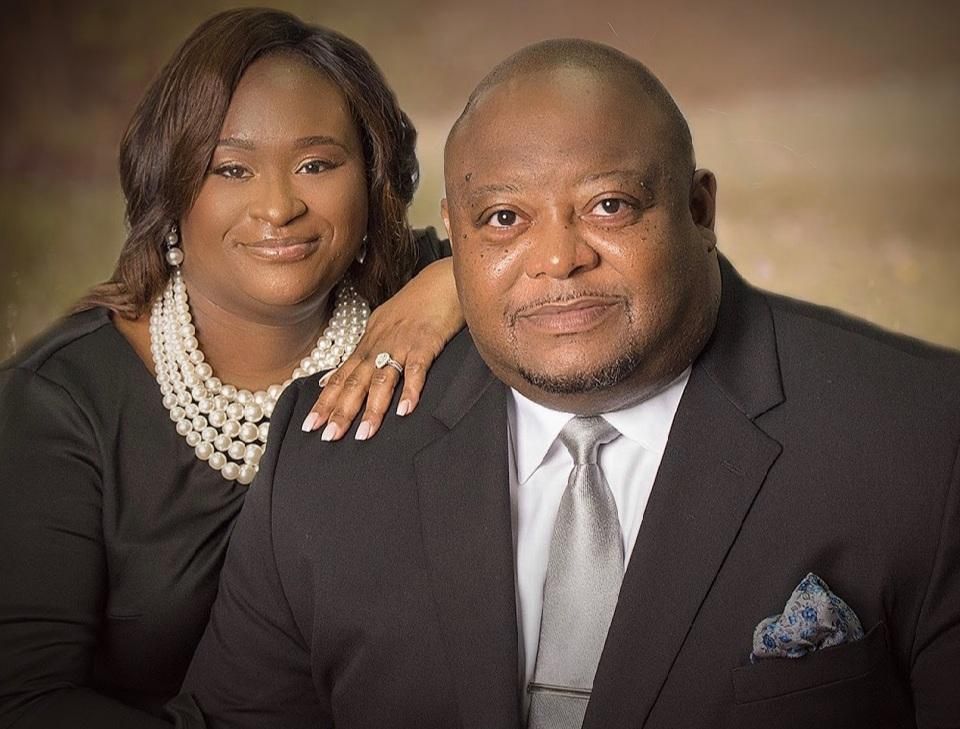 Couple posing, woman's hand on man's shoulder. Both are formally dressed. Soft background.