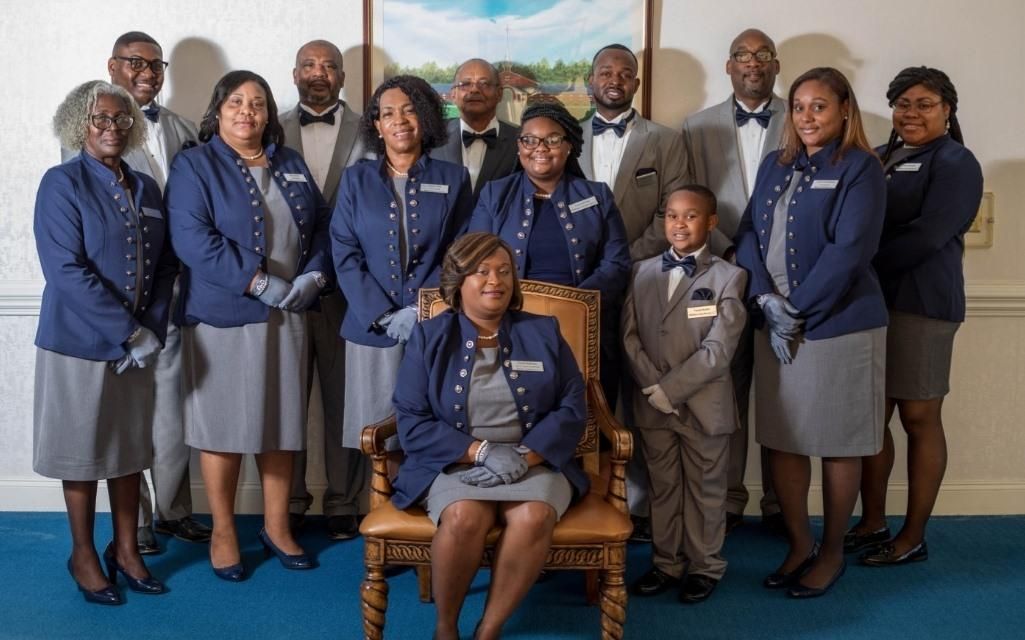 Group portrait: People in formal blue/gray attire in front of a painting; one person seated in a chair.