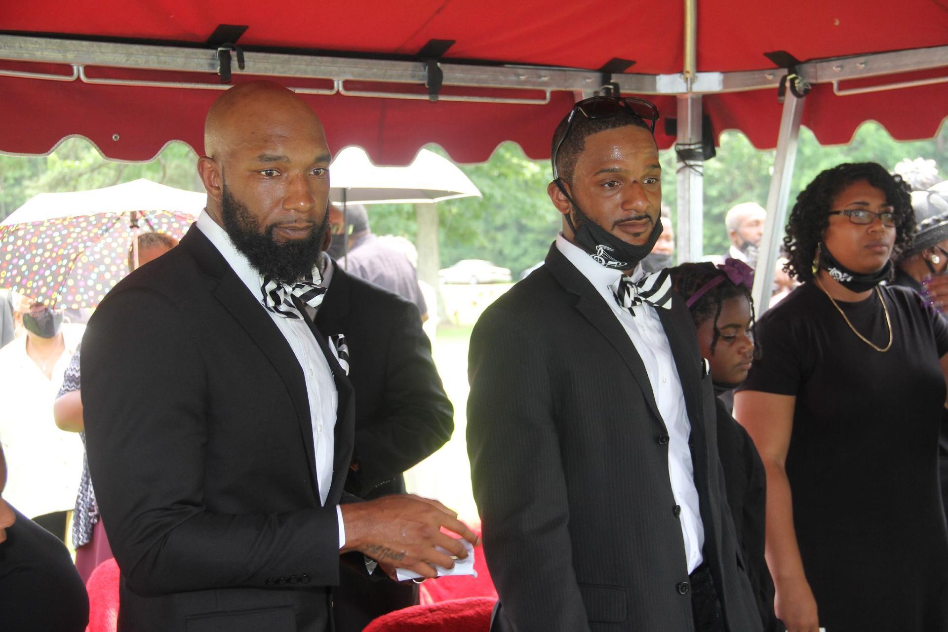 Two men in black suits and bow ties stand at an outdoor event.