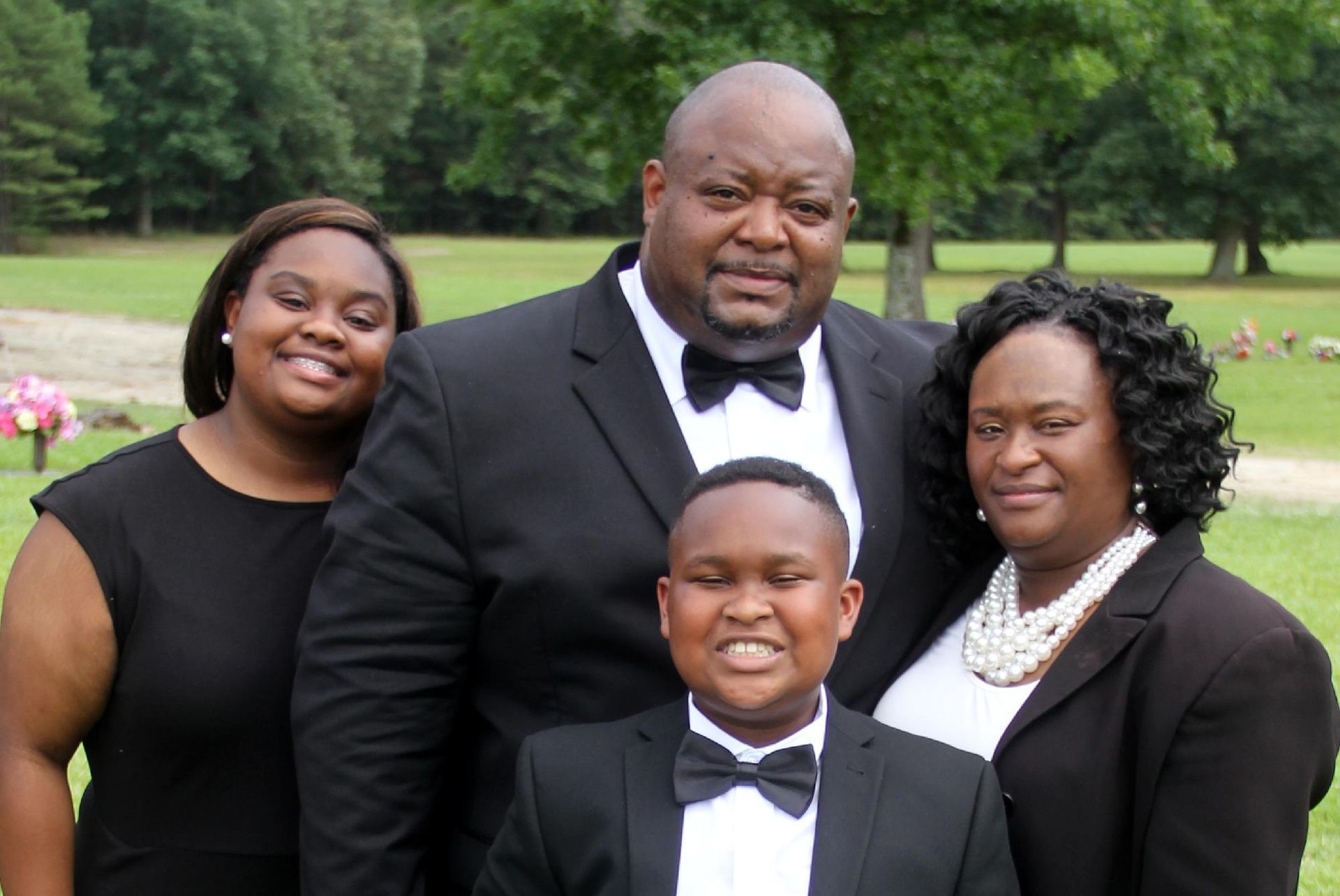 Family posing outdoors: two adults and two children in formal attire, smiling against a green background.