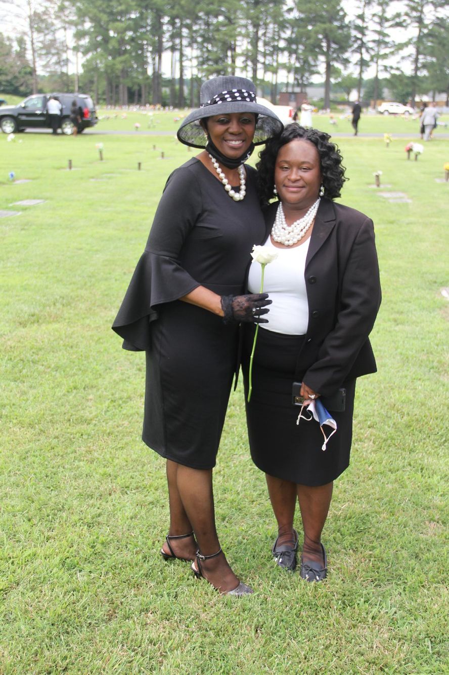 Two women pose outdoors, in a grassy cemetery. One wears a hat, the other holds a flower; both in black outfits.