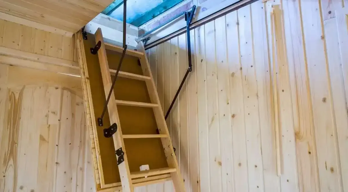 Wooden pull-down attic ladder extended from ceiling access in a wood-paneled room.