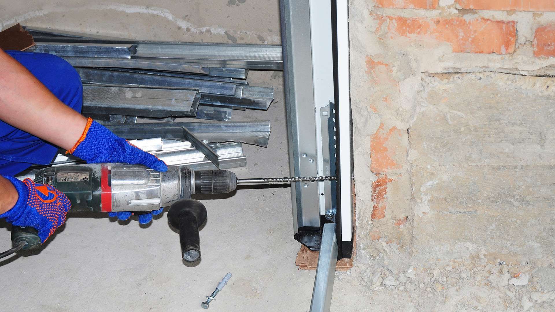 Person in blue overalls and gloves drilling into a metal door frame next to a brick wall.