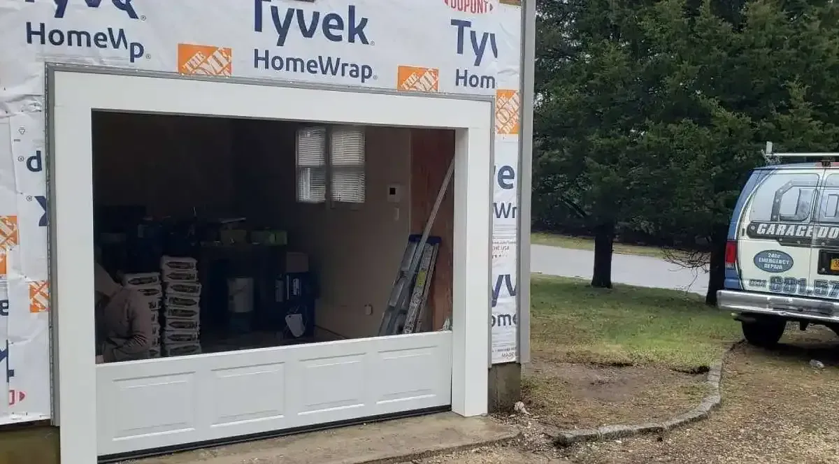 Garage door frame under construction with Tyvek house wrap. White frame, partially open. Van parked to the right.