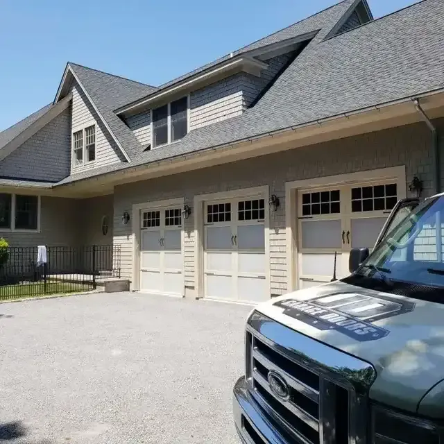 Three-car garage with beige doors, a gray roof, and a gravel driveway. A white truck is in the foreground.