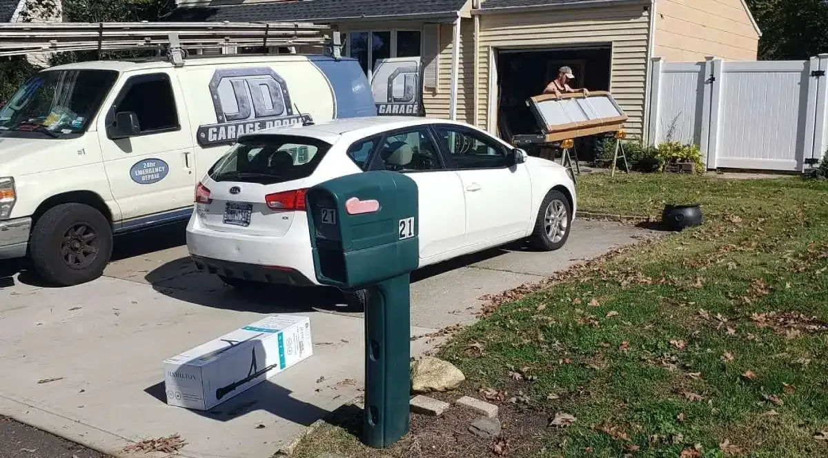 White car parked in driveway with a delivery box near a green mailbox. A work van and person in a garage.