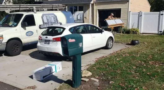 White car parked in driveway with a delivery box near a green mailbox. A work van and person in a garage.