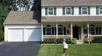 Two-story beige house with two-car garage, porch, green shutters, and a black driveway.