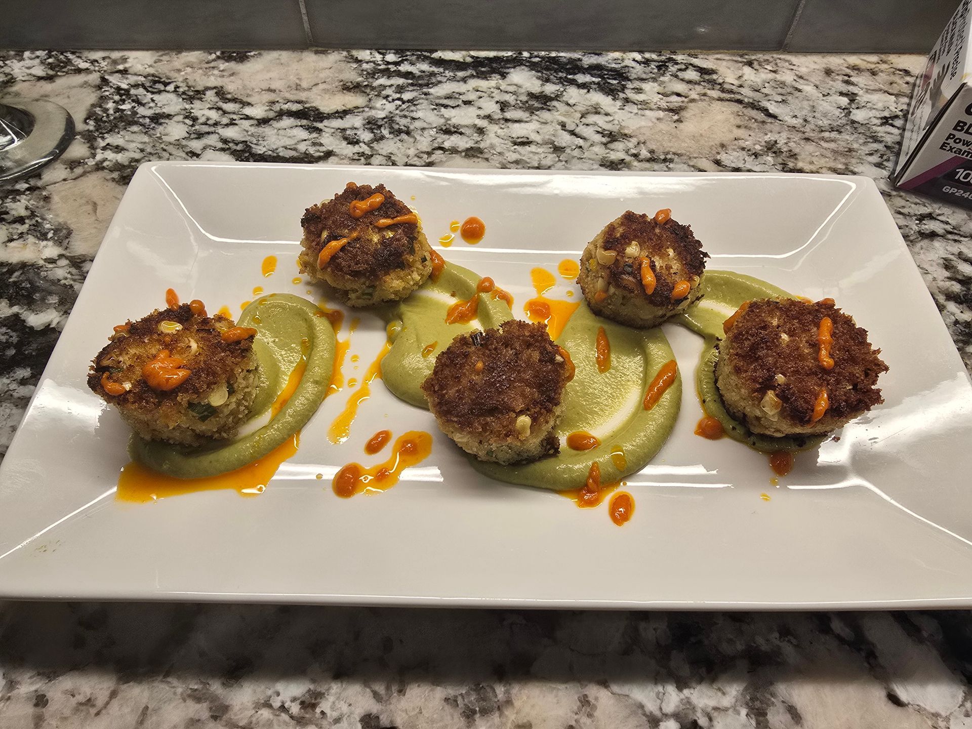 Six fried crab cakes on green puree, drizzled with orange sauce, on a white rectangular plate.
