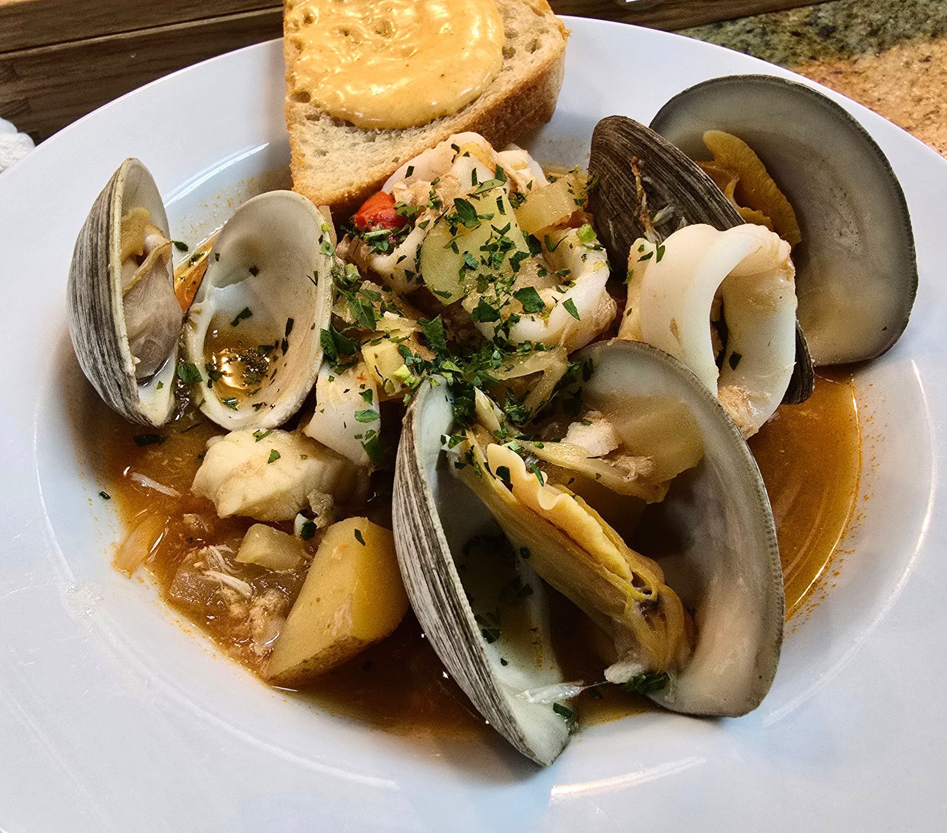 Seafood stew with clams, squid, and potatoes, served with toasted bread on a white plate.