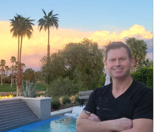 A man is standing in front of a pool with palm trees in the background