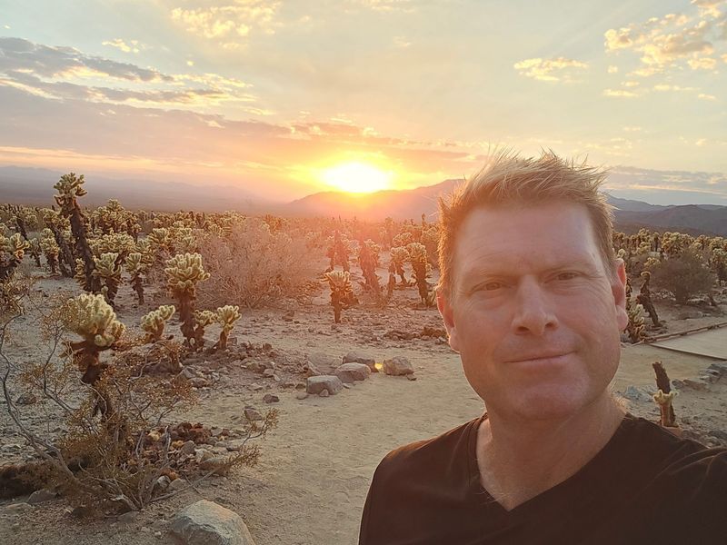 A man is taking a selfie in the desert at sunset.