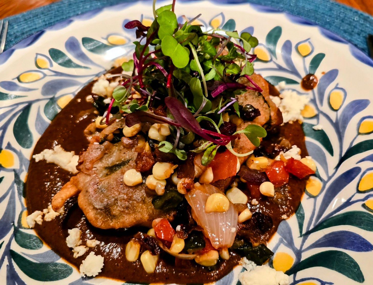 Dish of mole sauce topped with grilled vegetables, corn, cheese, and microgreens on a floral patterned plate.