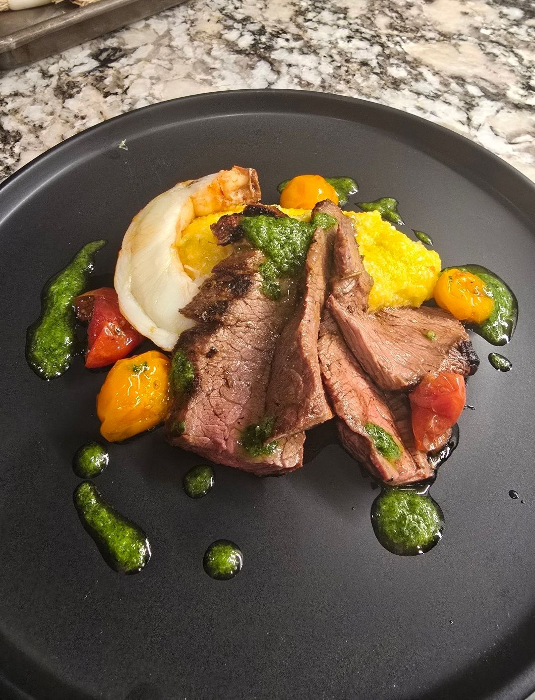 Steak and shrimp dish on black plate, with yellow and red tomatoes, green sauce.