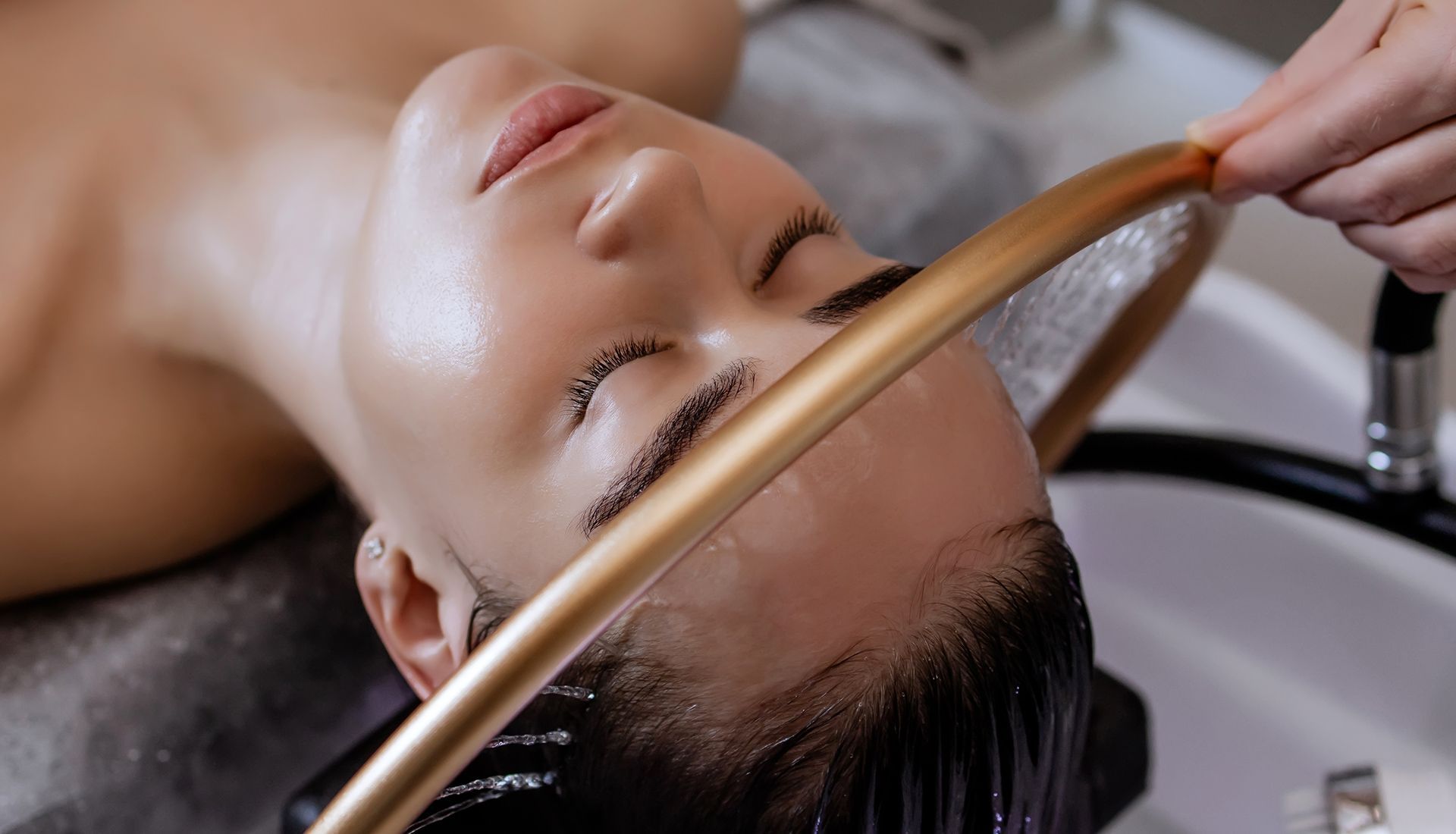 Person having hair washed at a salon. Water streams from a golden hose over their head into a sink.