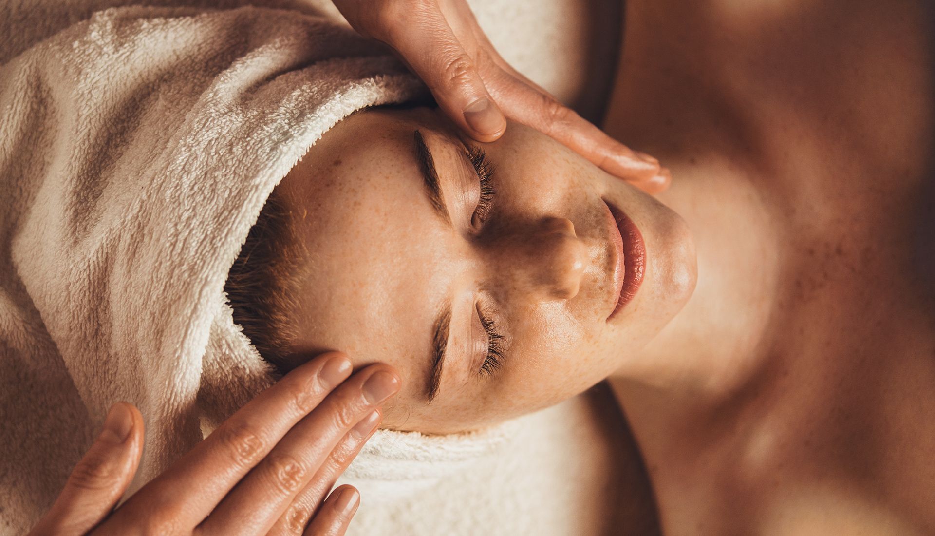 Woman receiving facial massage with closed eyes, head wrapped in towel.