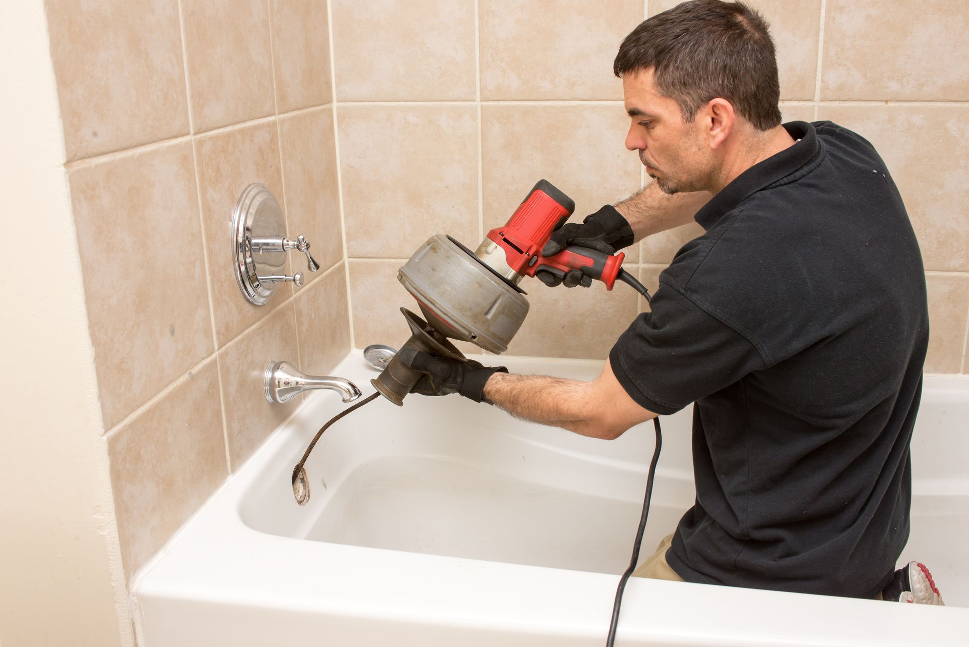 A technician from a plumbing company uses an electric snake tool to clear a bathtub.