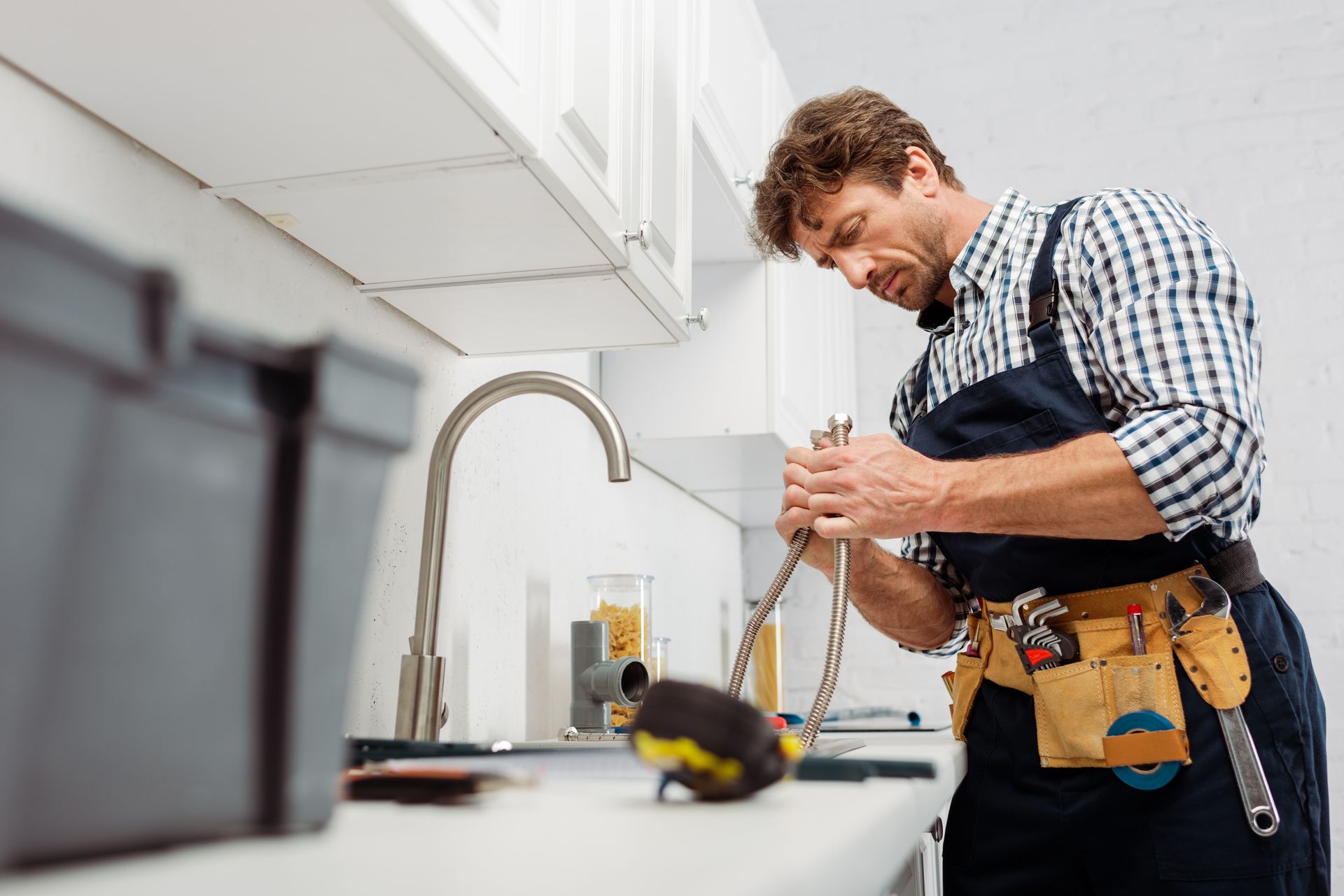 A plumber working at a kitchen sink, tightening a flexible water hose with tools on the counter