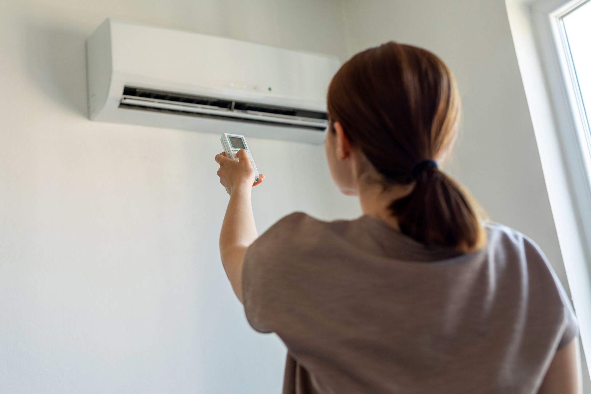 The backside of a woman turning on an air conditioner with a remote.