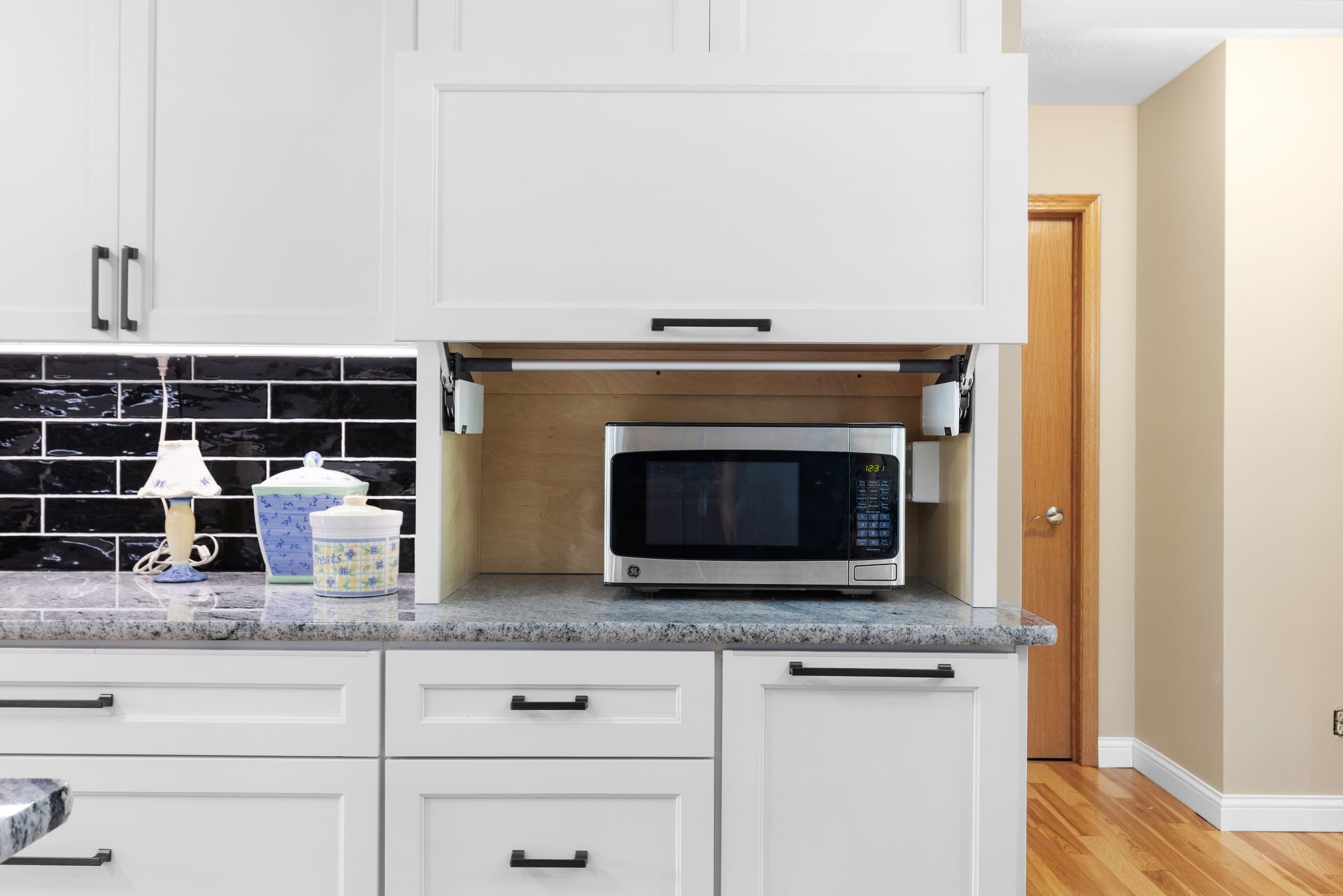 White kitchen cabinet with built-in microwave, door open. Light beige walls, wood floor, black tile backsplash.