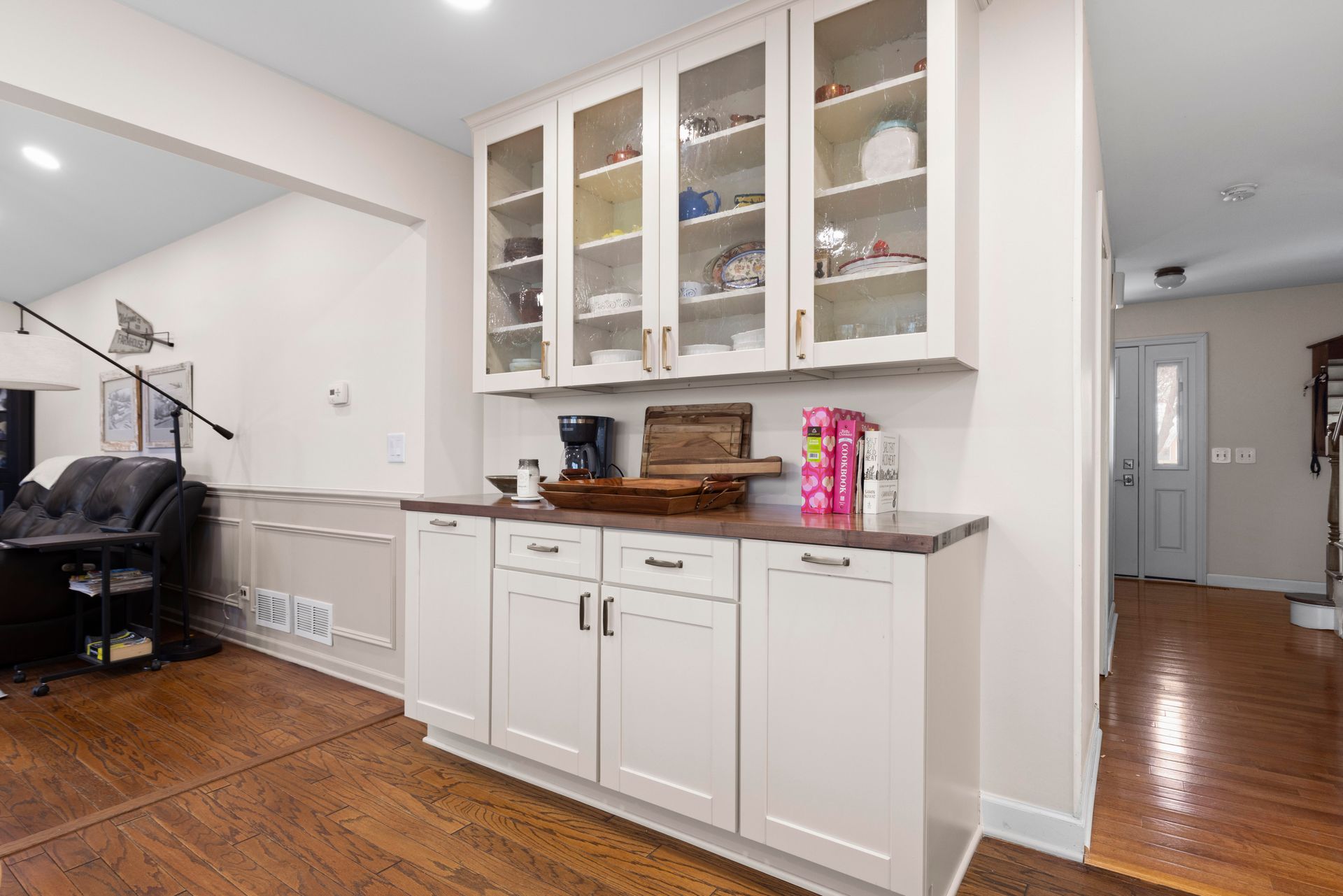 White kitchen cabinet with glass doors, housing dishware and coffee setup on countertop, hardwood floors.