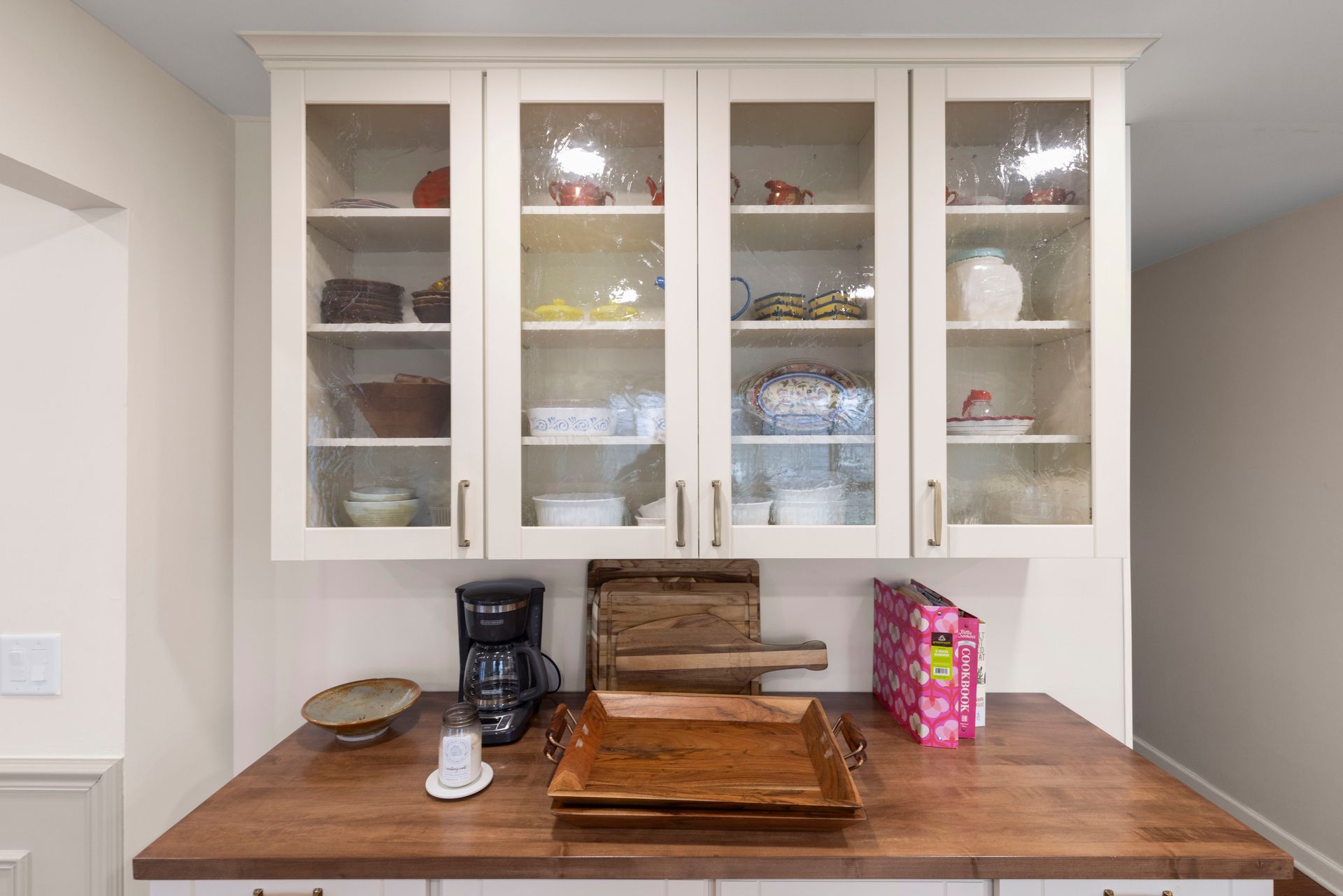 Kitchen cabinet with glass doors, above a countertop with coffee maker, trays, and decorative items.