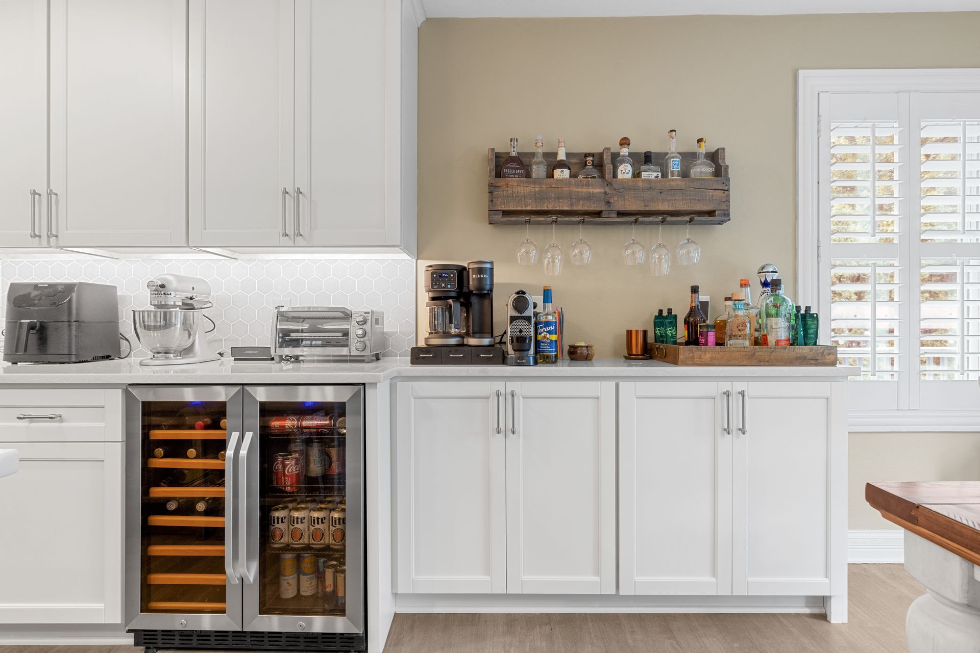 White kitchen cabinet with bar and wine cooler; bottles and glasses on display.