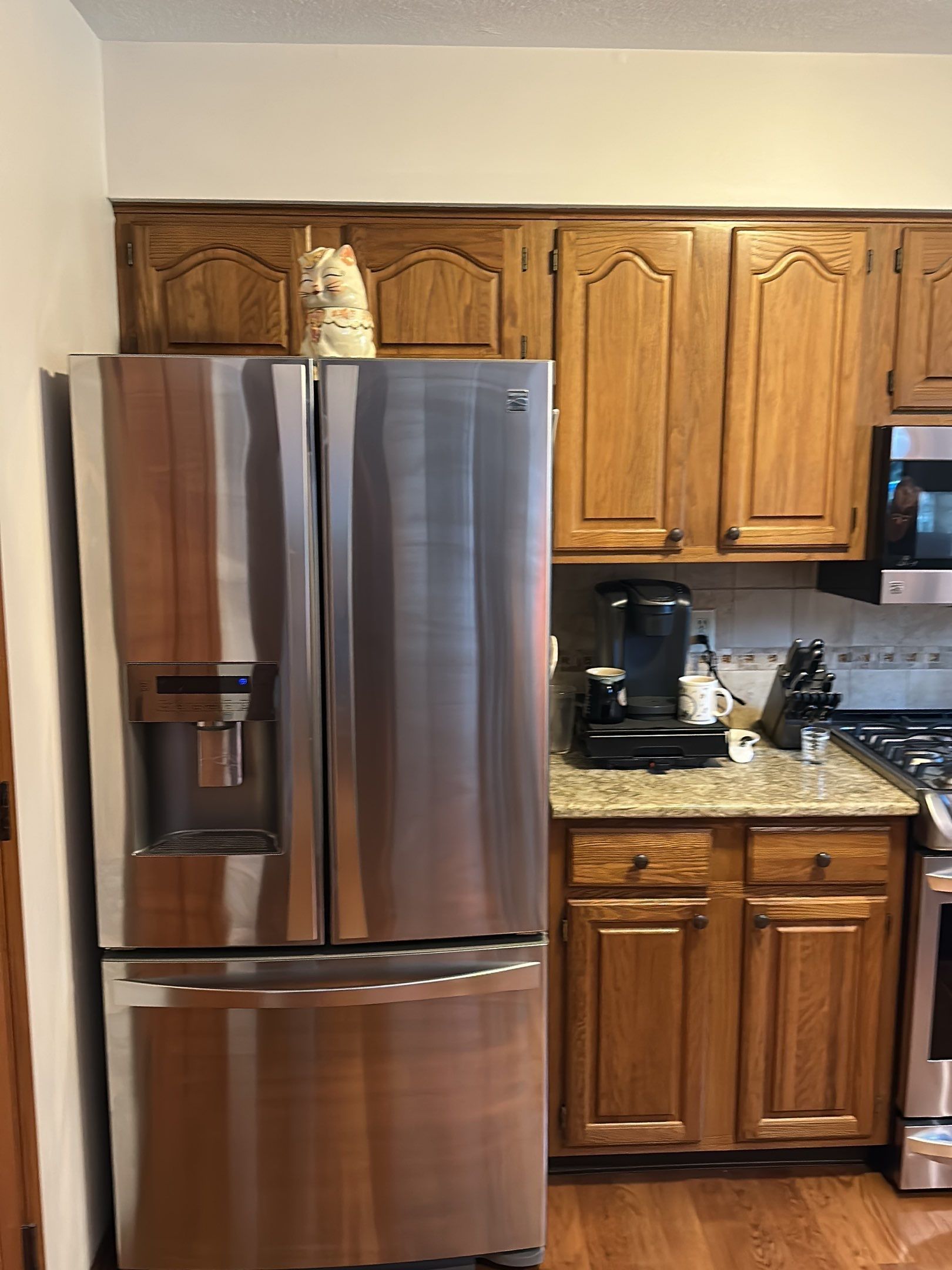Cat perched atop a stainless steel refrigerator in a kitchen with wooden cabinets and a coffee maker.