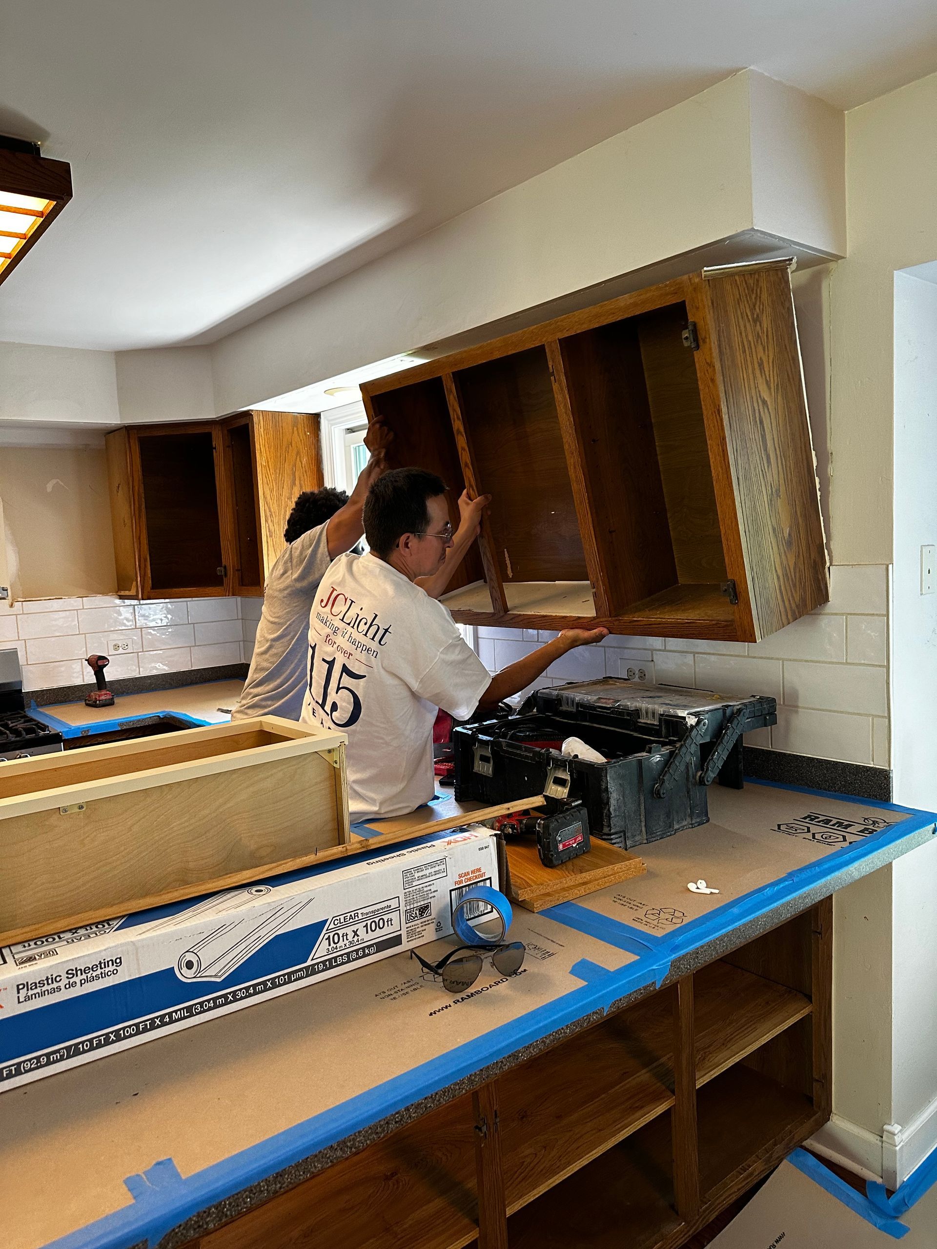 Two workers install a kitchen cabinet. Brown cabinets, white walls, and black tool box.