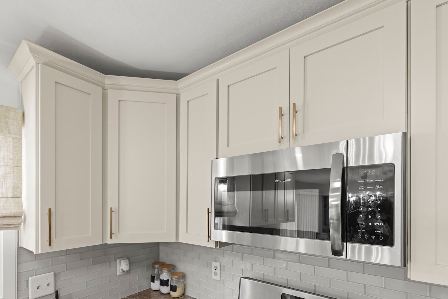Cream-colored kitchen cabinets with gold hardware, above a stainless steel microwave and gray backsplash.