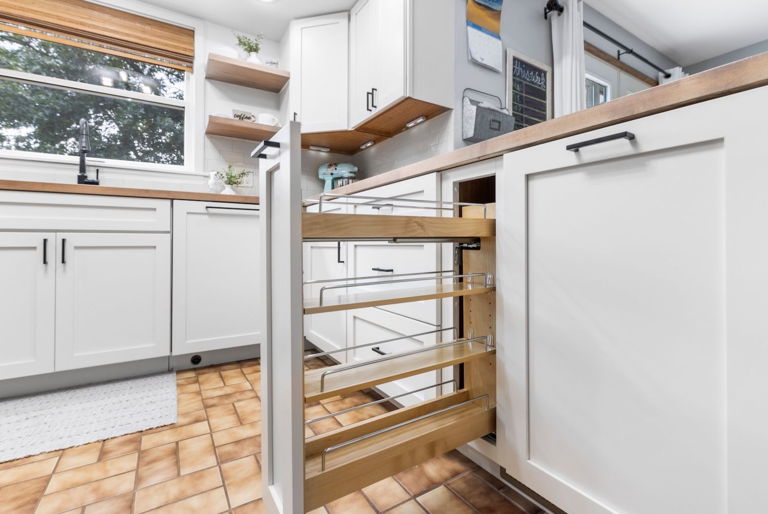 White kitchen with pull-out shelving, butcher block countertop, and a view of a window with blinds.