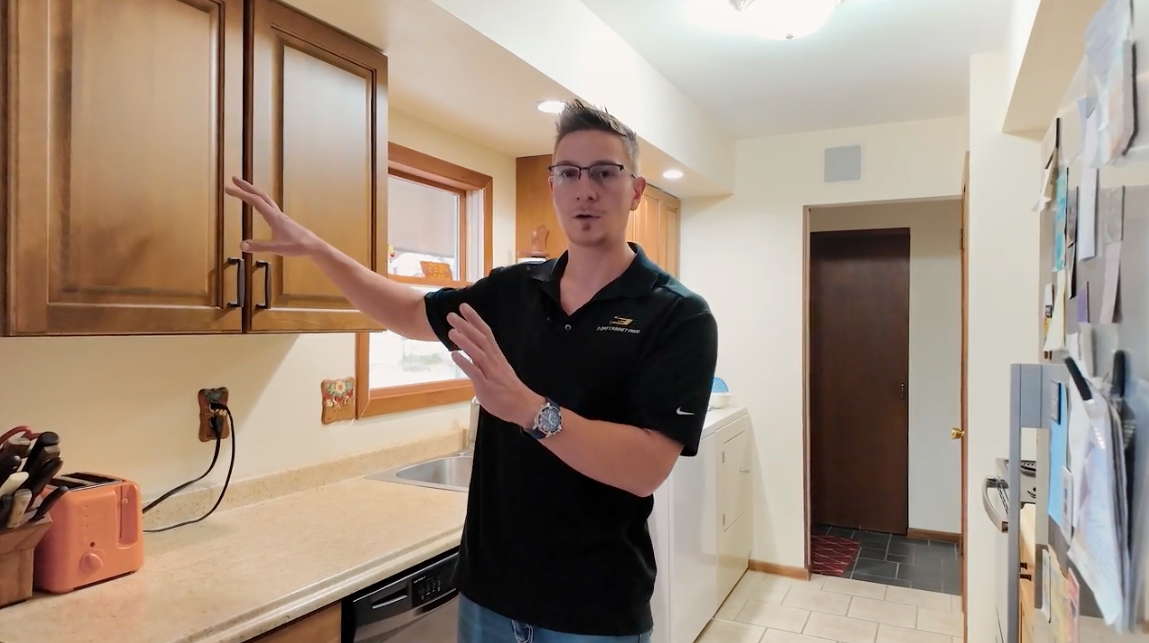 Man in kitchen gesturing towards upper cabinets; neutral tones, neutral expression.