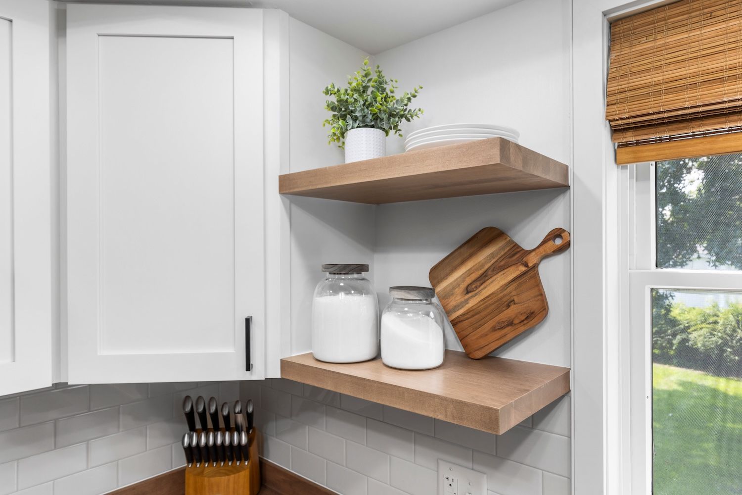 Kitchen corner with floating shelves holding jars, cutting board, and a plant. A window is to the right.