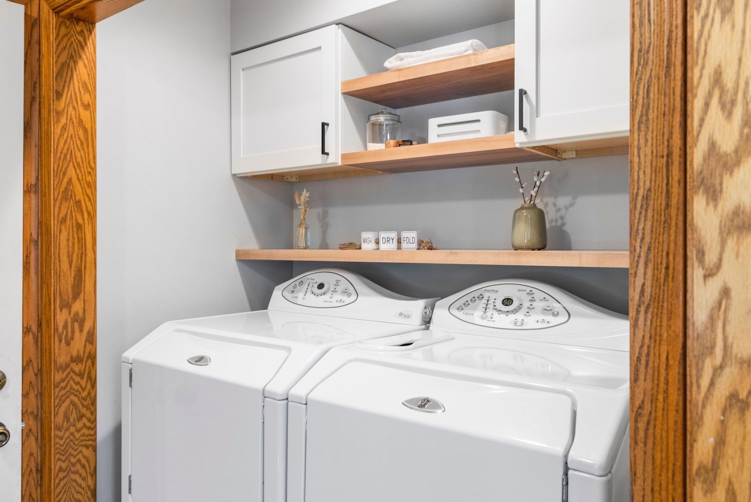 Laundry room with white washer/dryer, wooden shelves, white cabinets, and light gray walls.